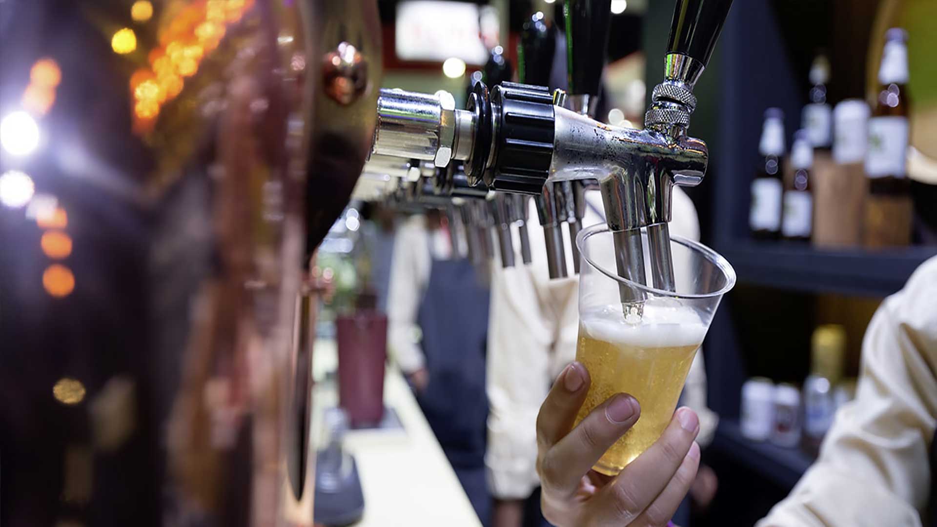Hand holding a clear plastic cup filled with beer under a beverage dispensing nozzle in a bar