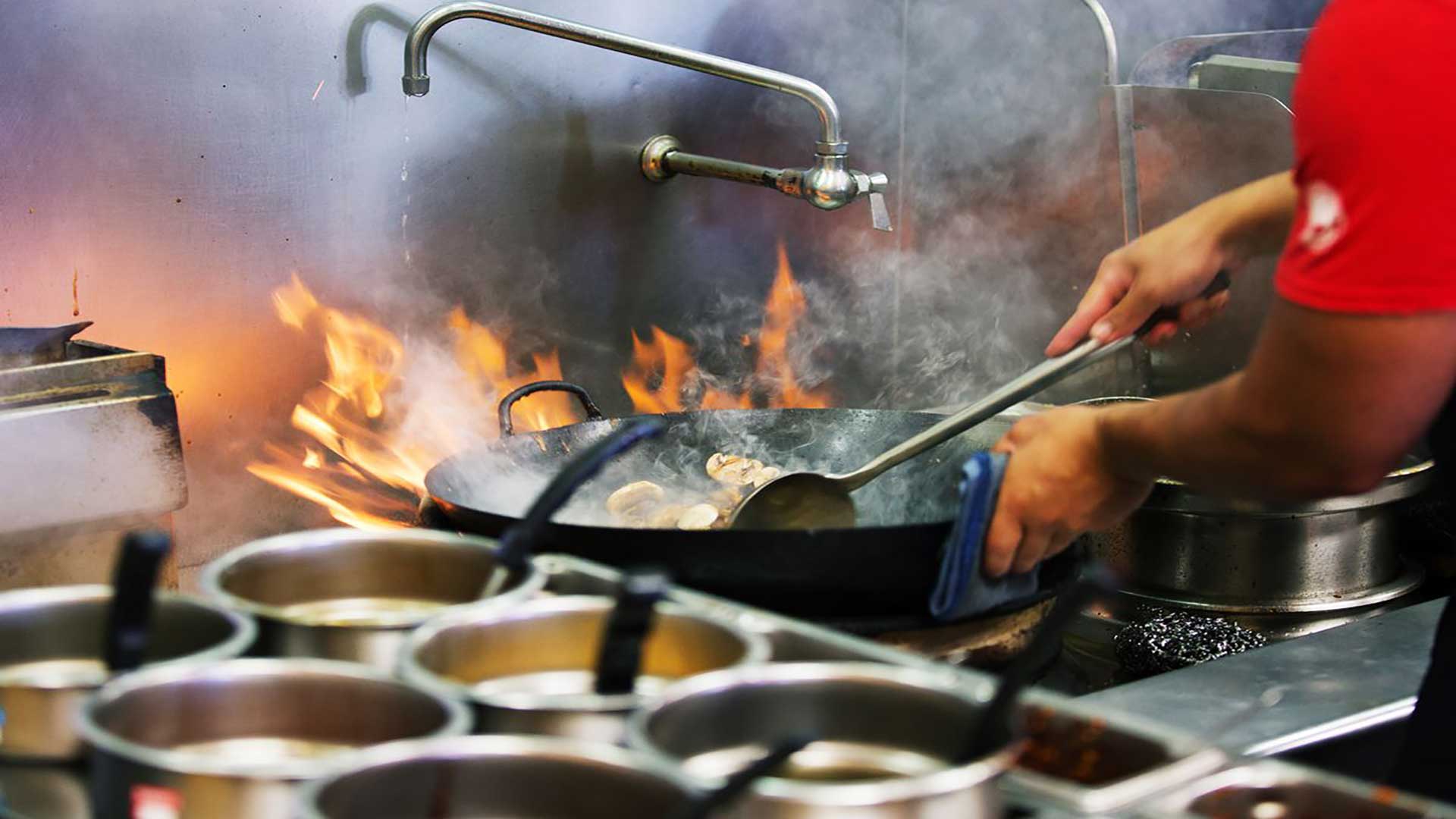 Person holding large wok above fire in a commercial kitchen