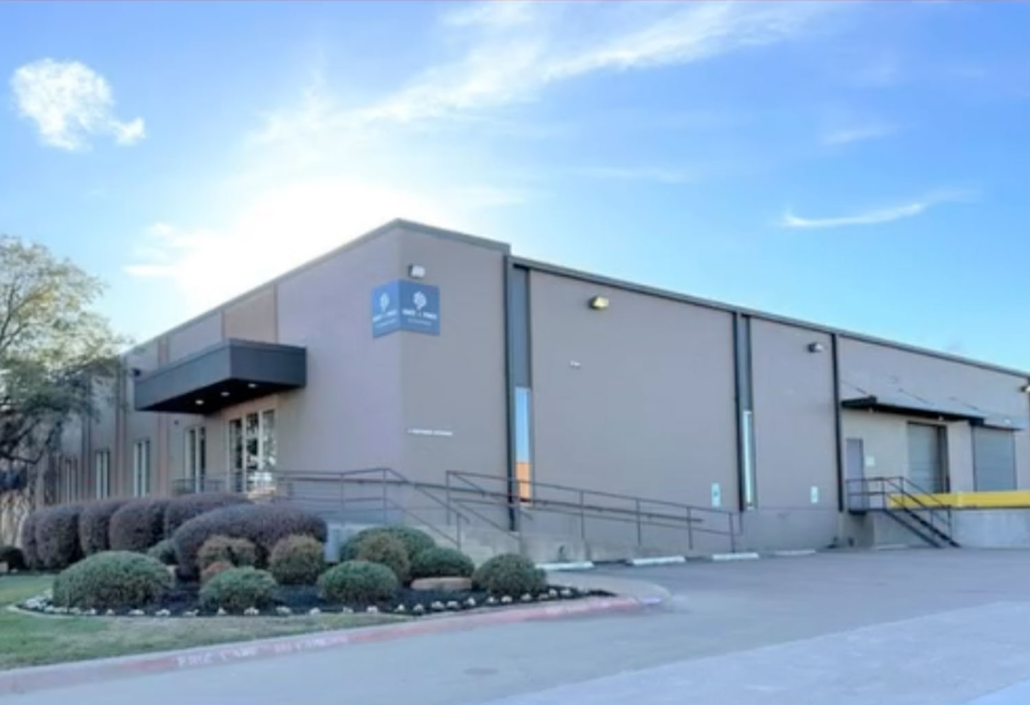 Hart-Price-Dallas-Texas-Location Hart+Price Dallas Texas location with a large, beige industrial building with a ramp, railings, and trimmed bushes at the entrance. Loading dock doors are on the side, and a blue sky with sun and clouds forms the backdrop. For more details, contact us today.