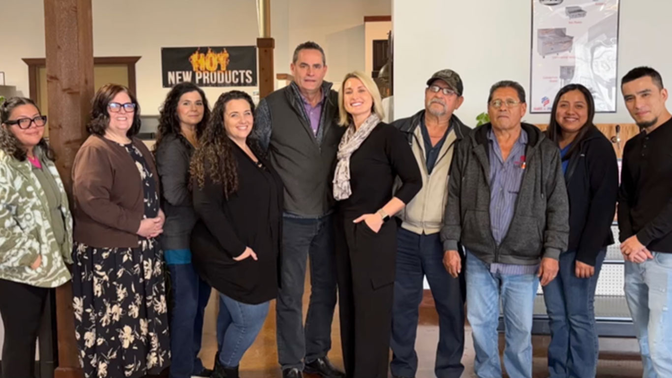 Hart-Price-Owners-and-Employees-Banner Owners and employees from Hart Price Foodservice Solutions stands indoors, smiling at the camera in casual and business-casual attire. They’re gathered closely in front of a "New Products" sign on the wall.