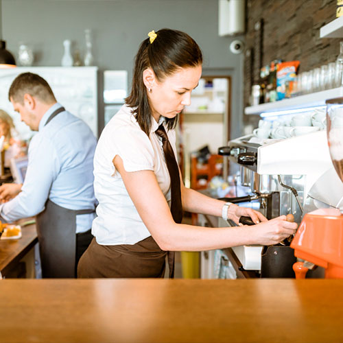 A woman in a white shirt and brown apron makes coffee using an espresso machine in a café, while a man in a blue shirt works in the background.
